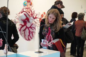 People in a gallery with a woman in the foreground looking at a soft sculpture of a human heart. Installation view from the exhibition 'Ema Shin, 'Hearts of Absent Women', Ararat Gallery TAMA, July 2023. Art Analog Club