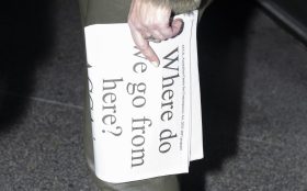 A pale hand holding a folded newspaper held vertically with the text ‘Where do we go from here?’
