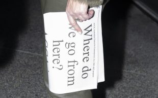 A pale hand holding a folded newspaper held vertically with the text ‘Where do we go from here?’