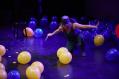 A woman blindfolded and squatting with a knife. There are yellow and blue balloons around her.