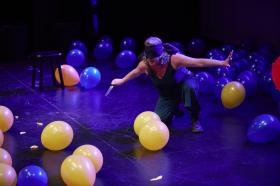 A woman blindfolded and squatting with a knife. There are yellow and blue balloons around her.