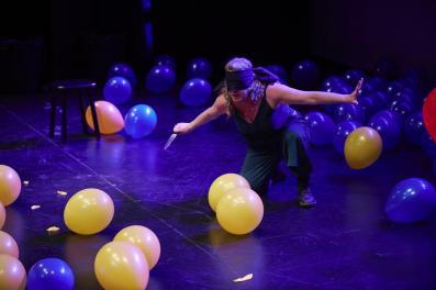 A woman blindfolded and squatting with a knife. There are yellow and blue balloons around her.