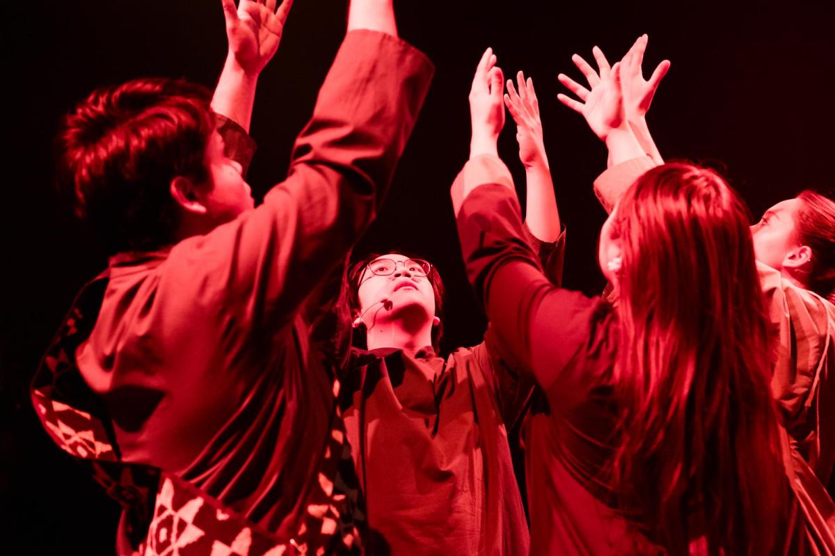 Four performers bathed in red light have their hands in the air. They are in a huddle. Scenes of a Yellow Peril.