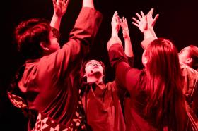 Four performers bathed in red light have their hands in the air. They are in a huddle. Scenes of a Yellow Peril.