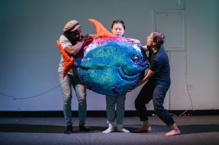 Three people on stage with a blue fish puppet. Creative Futures Fund