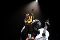 Singer-songwriter Ed Sheeran, a fair-skinned man with red hair, plays an accoustic guitar live on stage while standing in front of a screen showing a larger-than-life black and white image of himself.