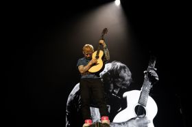 Singer-songwriter Ed Sheeran, a fair-skinned man with red hair, plays an accoustic guitar live on stage while standing in front of a screen showing a larger-than-life black and white image of himself.