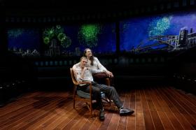 A man is sitting on a chair and a woman is standing beside him. The view of Sydney Opera House can be seen behind them.