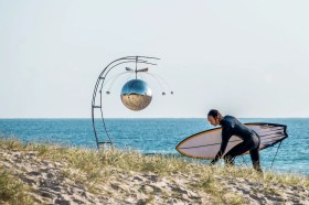 Man in wetsuit with surfboard on beach with sculpture. SWELL Sculpture Festival