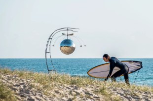 Man in wetsuit with surfboard on beach with sculpture. SWELL Sculpture Festival