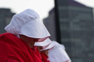 From the side we see a woman in a red cloak and white bonnet who bows her head so we can't see her face; she is in a line with other women dressed the same. The Handmaid's Tale.