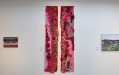 A textile work of two thin strips in red, pink, brown and light purple, hung on a white wall.