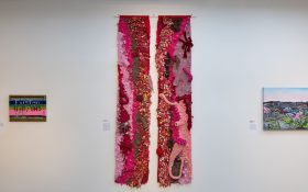 A textile work of two thin strips in red, pink, brown and light purple, hung on a white wall.