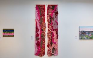 A textile work of two thin strips in red, pink, brown and light purple, hung on a white wall.