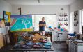 Ken Done, an 85-year-old Caucasian man standing smiling with his arms crossed on top of his chest inside a studio with canvas, paint, and a work in progress.