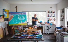 Ken Done, an 85-year-old Caucasian man standing smiling with his arms crossed on top of his chest inside a studio with canvas, paint, and a work in progress.