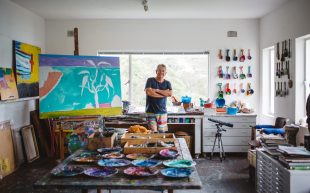 Ken Done, an 85-year-old Caucasian man standing smiling with his arms crossed on top of his chest inside a studio with canvas, paint, and a work in progress.