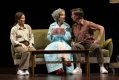 Three people sit on a couch, a mousy woman in her 30/40s dressed in brown, an older woman in a turquoise housecoat and turban and a man in his 30/40s dressed in brown and facing off with the older woman. A coffee table is in front of them. Mother Play.