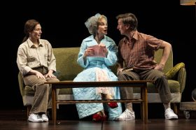 Three people sit on a couch, a mousy woman in her 30/40s dressed in brown, an older woman in a turquoise housecoat and turban and a man in his 30/40s dressed in brown and facing off with the older woman. A coffee table is in front of them. Mother Play.