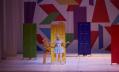 Awoman in a blue dress is standing in front of a yellow door. A person in yellow is next to her. The backdrop is made of colourful shapes.