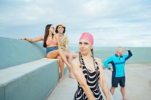 A woman with grey plaits escaping from under a pink bathing cap stares at the camera. Behind her, sitting on the steps of the Newcastle Ocean Baths or standing nearby, are three other actors dressed for a swim or the beach. The ocean and sky meet behind them, in this publicity photo for 'Meet Me at the Baths' at New Annual Festival.