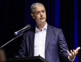 NSW Minister for the Arts John Graham MLC launches the Plan for Western Sydney Arts, Culture and Creative Industries: 2025-2028. The photo shows an animated man with silver hair and wearing a blue suit and white shirt but no tie. He stands at a lectern and gestures with his left hand.