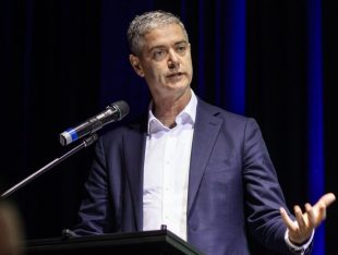 NSW Minister for the Arts John Graham MLC launches the Plan for Western Sydney Arts, Culture and Creative Industries: 2025-2028. The photo shows an animated man with silver hair and wearing a blue suit and white shirt but no tie. He stands at a lectern and gestures with his left hand.