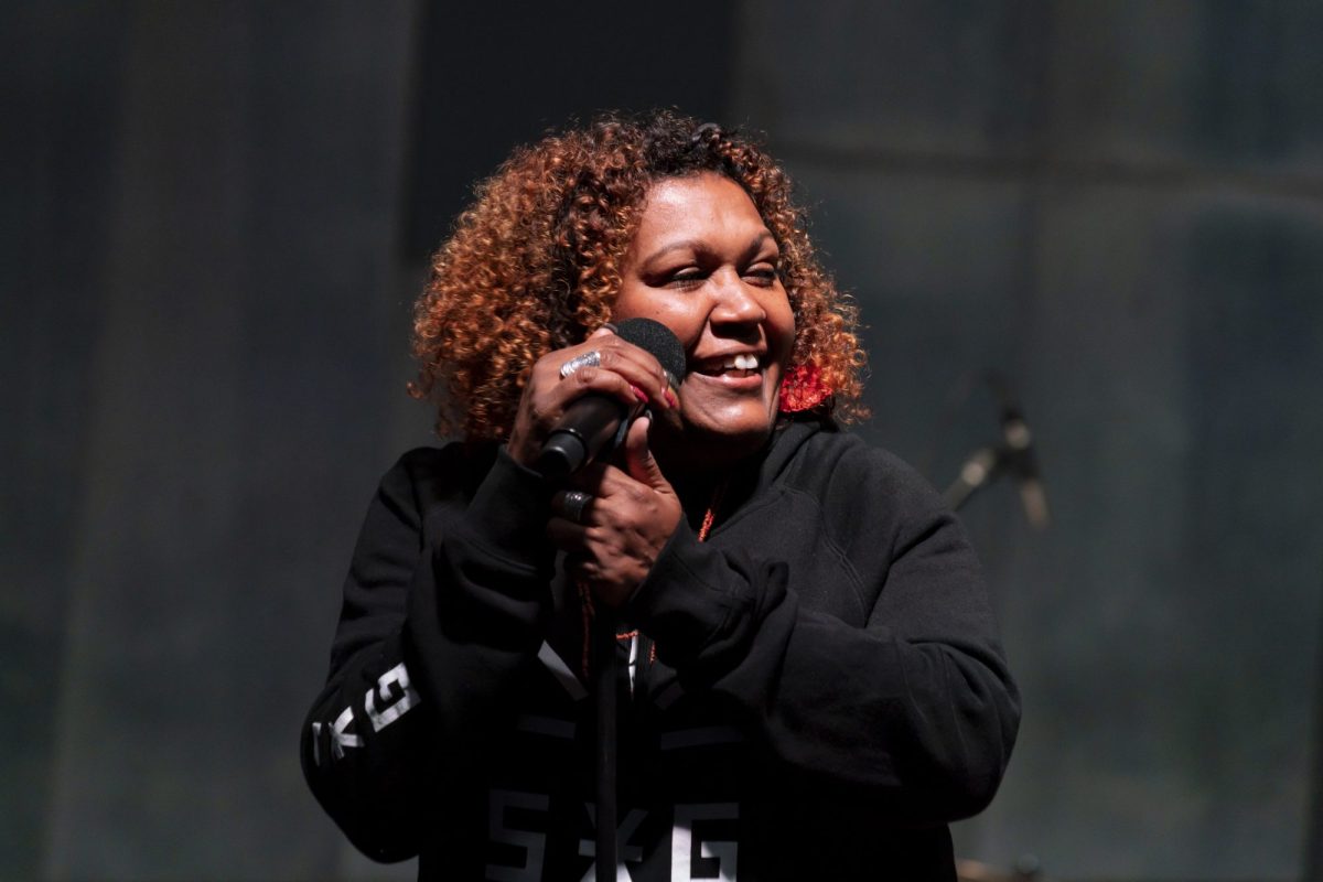 a First Nations woman in a black shirt with white writing on it holds a microphone and smiles off to the side. Emma Donovan. Port Fairy Folk Festival. NAIDOC in the City