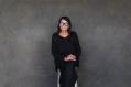 A woman in black with blue glasses sitting against a grey wall. Performer of 'My Cousin Frank'.
