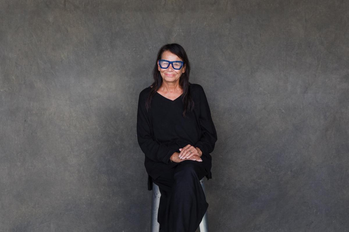 A woman in black with blue glasses sitting against a grey wall. Performer of 'My Cousin Frank'.