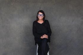 A woman in black with blue glasses sitting against a grey wall. Performer of 'My Cousin Frank'.
