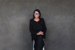 A woman in black with blue glasses sitting against a grey wall. Performer of 'My Cousin Frank'.