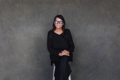 A woman in black with blue glasses sitting against a grey wall. Performer of 'My Cousin Frank'.