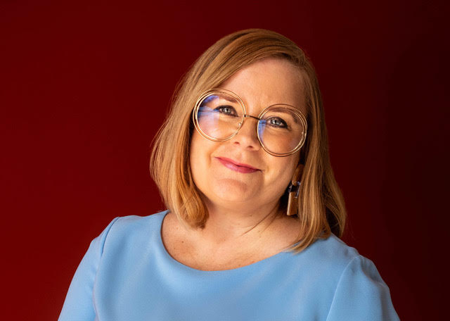 a woman in a light blue top with large glasses and a light brown bob smiles at the camera with her head leaning slightly to one side. Natalie Jenkins.