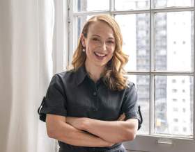 Olivia Ansell: a portrait photograph of a smiling white woman in her forties with long light brown hair with her arms crossed in front of her, wearing a black short sleeved top standing in a light-filled white-walled room next to a window with light coming in through the window. Toronto