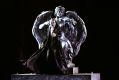 A sinister figure wearing a half-mask over his face and 19th Century clothes, including a long, black, cloak-like coat, gestures dramatically as he sings. He stands in front of a large statue of an angel playing the violin in the scene from Opera Australia's Handa Opera on Sydney Harbour 2022 production, 'Phantom of the Opera'.
