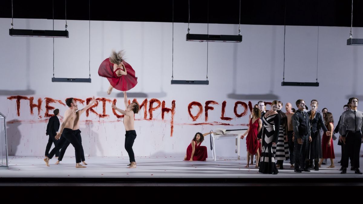 a group of performers including some bare-chested circus performers on stage in a scene from Opera Queensland and Circa's world premiere production of 'Orpheus & Eurydice', 2019. The back white wall has THE TRIUMPH OF LOVE in red scrawled across it.