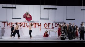 a group of performers including some bare-chested circus performers on stage in a scene from Opera Queensland and Circa's world premiere production of 'Orpheus & Eurydice', 2019. The back white wall has THE TRIUMPH OF LOVE in red scrawled across it.