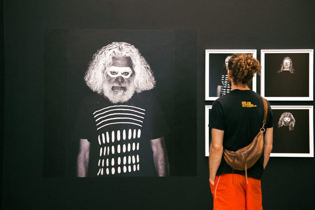 Person wearing orange pants and black T shirt looking at First Nations work on a black wall - a photo of a man with white hair, a black and white t shirt and white paint around his eyes, and four small photos. ‘On Country: Photography from Australia’, Rencontres d'Arles 2025.