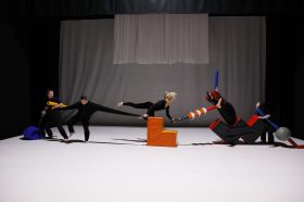 A production photo of dynamic dancers dressed in black suits, forming single file while pulling at each other’s clothes. RISING