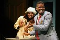 A black woman and a black man are laughing. She's wearing a white hat and a yellow suit, he's wearing a grey suit with a red tie.