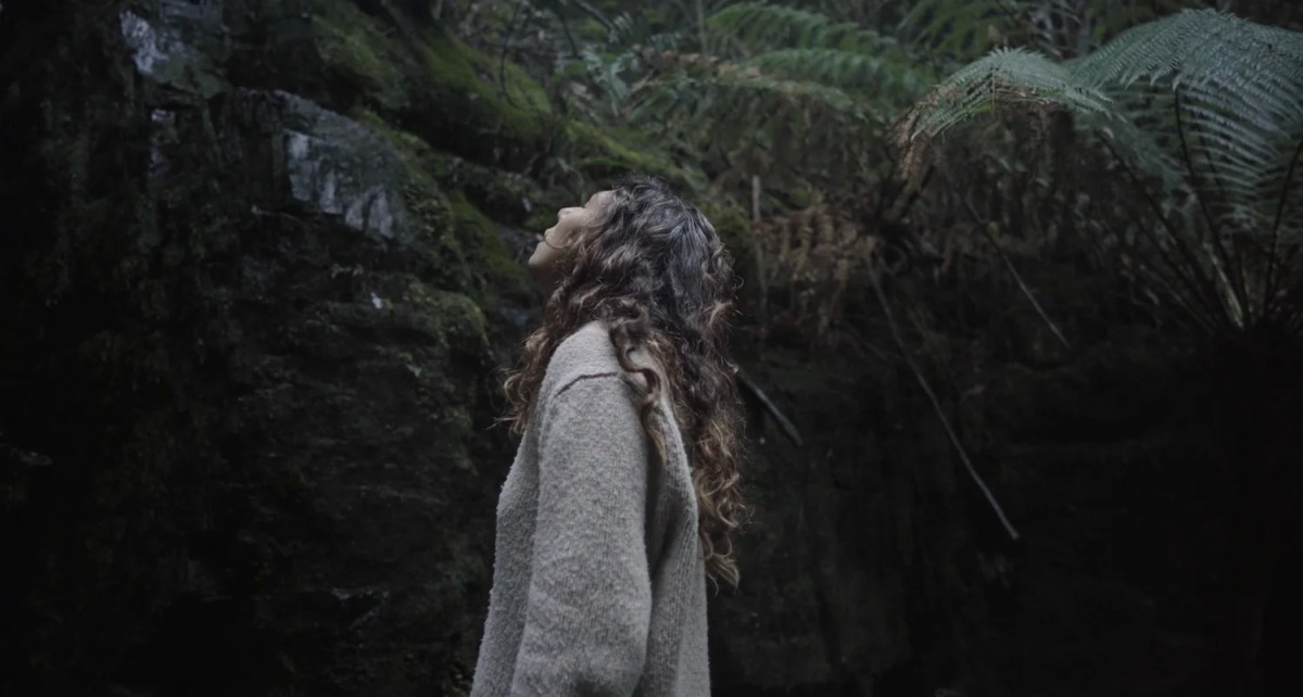 A woman looks up a dark cliff face, an example of pleasure pilgrimages in Tasmania.
