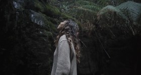A woman looks up a dark cliff face, an example of pleasure pilgrimages in Tasmania.