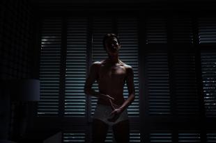 A man wearing white underwear is standing in the dark in front of some blinds. American Psycho.