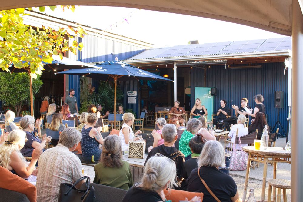 Audience members applaud panellists at Regional Arts Development Organisation South Coast Arts' creative mixer in 2025. The photo shows a group of five female-presenting people seated on a low stage, with two dozen or more people seated in a courtyard and applauding them.