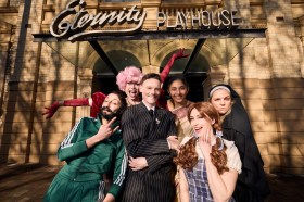 A man in a black suit surrounded by five other people in different costumes.