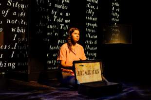 The 7 Stages of Grieving: A young Aboriginal female performer kneels onstage against a black backdrop that has cursive white text written on it displaying words like Nation', 'Telling' and 'People'. She is holding a small sign that says 'reconciliation'.