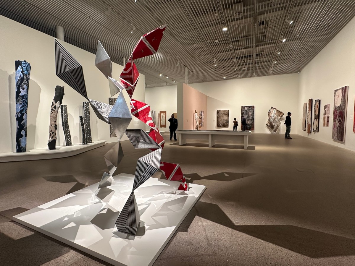 Gallery setting with Aboriginal artworks on display with a bit metal sculpture in the foreground. Yirrkala