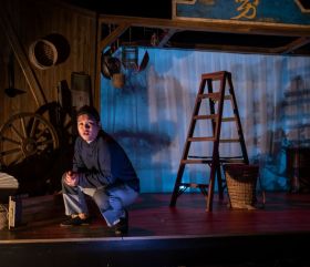 A young boy is crouching to the side. Behind him is a ladder in a production of Yong.