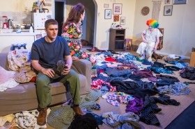 A man sitting on a couch, a woman behind him. In the background on the right is a man dressed like a clown. On the ground are clothes strewn everywhere. Hir.